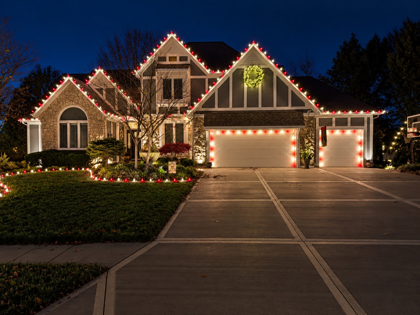 Two-story stone and siding home near Gardner, Kansas lit for Christmas: alternating red and warm white bulbs along every gable and roofline, both garage doors outlined to match, a lit wreath on the front gable, and red and white stake lights edging the front beds
