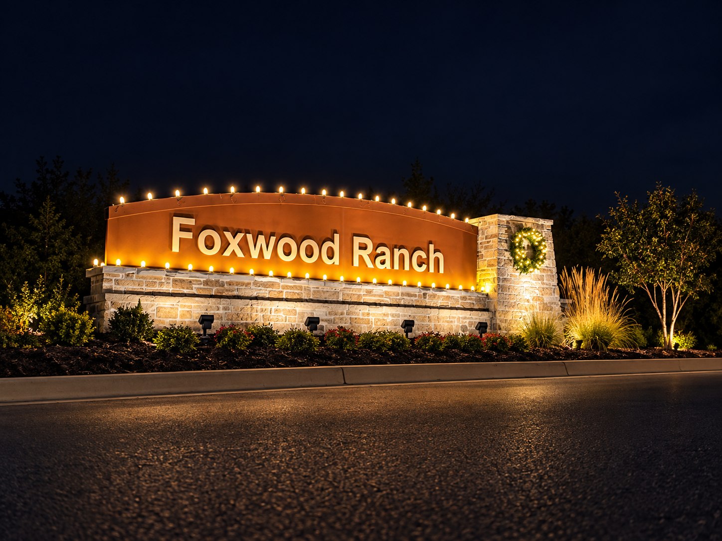 Foxwood Ranch neighborhood entrance sign at night, lined with warm white bulbs, with a lit wreath on its stone pillar